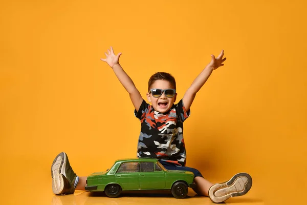 Dziecko w okularach przeciwsłonecznych, koszulce z kamuflażem. Podniesione ręce, siedzące na podłodze w pobliżu zielonego modelu samochodu retro, pozujące na pomarańczowym tle — Zdjęcie stockowe