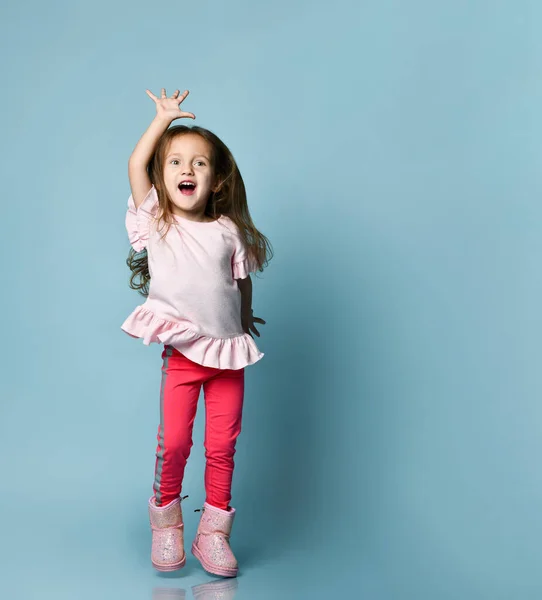 Klein blond meisje gekleed in roze blouse, broek en laarzen. Ze poseert tegen een blauwe studio achtergrond. Volledige lengte, kopieerruimte — Stockfoto