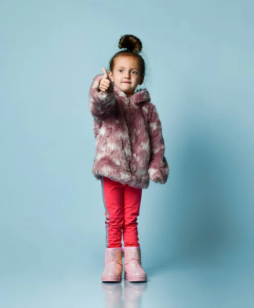 Klein meisje gekleed in roze nepbontjas, broek en laarzen. Ze glimlacht, wijst naar jou, poseert op een blauwe achtergrond. Totale lengte — Stockfoto