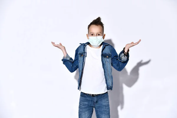 Blond teenagedreng i blå jeans, jakke, t-shirt. Han er i medicinsk maske, ser forvirret, forklædt isoleret på hvid. Coronavirus - Stock-foto