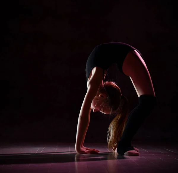 Giovane ginnasta ragazza in corpo nero sport e calzature speciali in piedi in posa ponte e mantenere l'equilibrio su sfondo scuro — Foto Stock