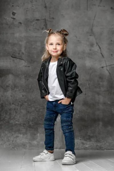 Little smiling blond girl in stylish rock style black leather jacket, blue jeans and white sneakers standing with hands in pockets Royalty Free Stock Images