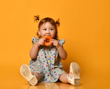 İki at kuyruklu, renkli elbiseli ve beyaz sandaletli küçük kız. Yerde oturmak, donut yemek, turuncu arka planda poz vermek.