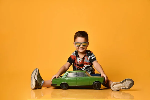 Kid in sunglasses, camouflage t-shirt, sneakers. Sitting on floor, holding green model of retro car, posing on orange background