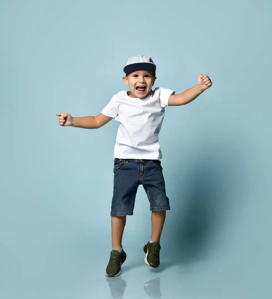 Bambino in t-shirt bianca, berretto grigio, pantaloncini di jeans e scarpe da ginnastica kaki. Saltò su con la bocca aperta, posando su sfondo blu — Foto Stock