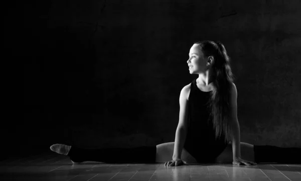 Young smiling girl gymnast in black sport body and uppers sitting in twine on floor and holding pink gymnastic ball in hands — ストック写真