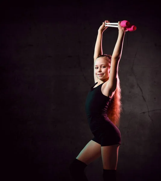 Giovane ragazza sorridente ginnasta in corpo nero sport e tomaie in piedi e in possesso di due mazze da ginnastica rosa in mani in rilievo — Foto Stock