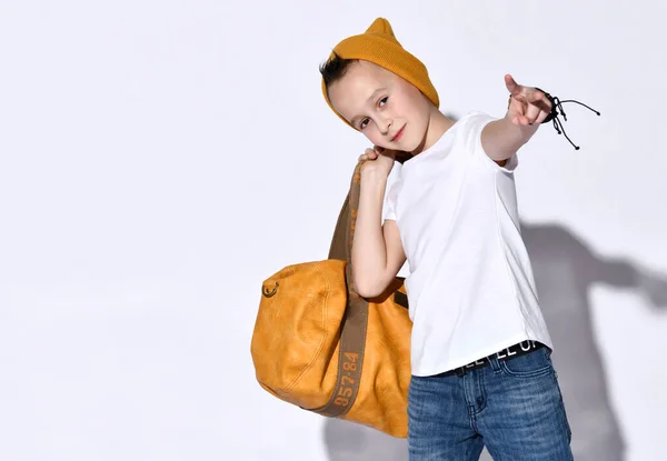 Ragazzo adolescente con borsa da viaggio arancione, in cappello, t-shirt, jeans blu, braccialetto. Sorridente, mostrando la mano della vittoria, posa isolata sul bianco — Foto Stock