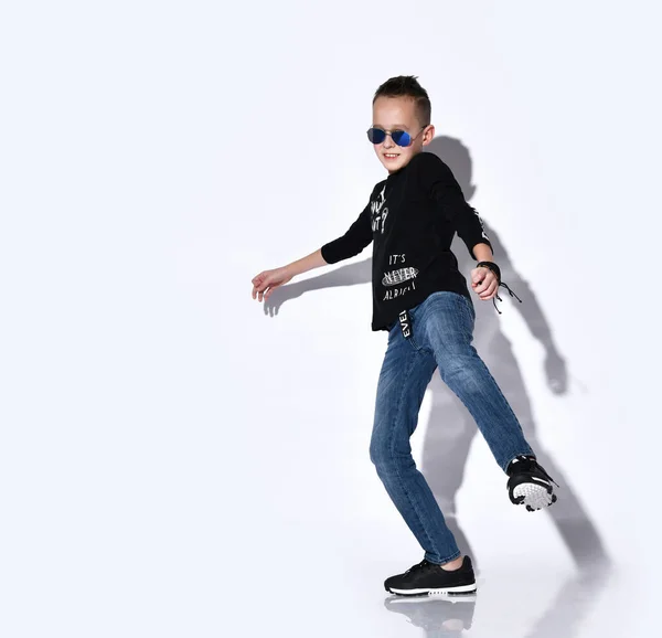 Niño adolescente en gafas de sol, jersey negro y zapatillas de deporte, vaqueros azules, pulsera. Bailando, posando aislado sobre fondo blanco del estudio —  Fotos de Stock