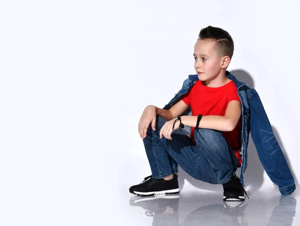 Bambino adolescente in t-shirt rossa, jeans e giacca in denim, sneakers nere. Indicando qualcosa, accovacciato lateralmente isolato su bianco — Foto Stock