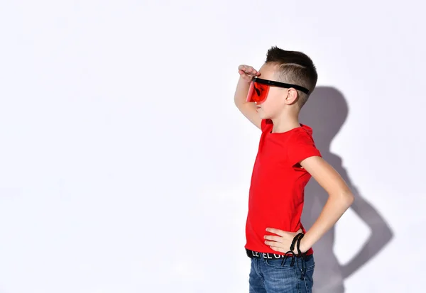 Nastolatek w wielkich okularach przeciwsłonecznych, czerwonym t-shircie, czarnych bransoletkach. Wyglądając na zdziwionego, wskazując na coś, udając odizolowanego na białym — Zdjęcie stockowe