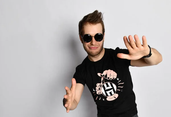 Hipster man i solglasögon, svart t-shirt med tryck, armband. Han ler och gestikulerar, poserar isolerad på vitt. Närbild — Stockfoto