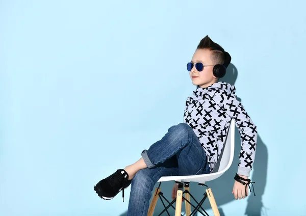 Tonårspojke i hörlurar, solglasögon, blå jeans, luvtröja, sneakers. Visar segertecken, sitter på vit stol. Blå bakgrund — Stockfoto