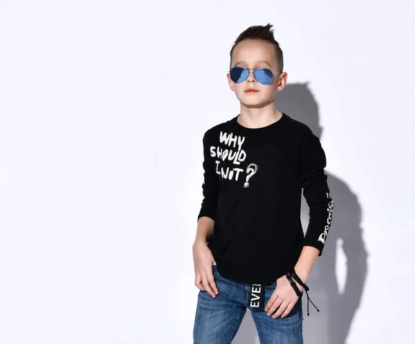 Niño adolescente en gafas de sol y jersey negro con inscripciones, vaqueros azules, pulsera. Posando aislado sobre fondo blanco del estudio — Foto de Stock