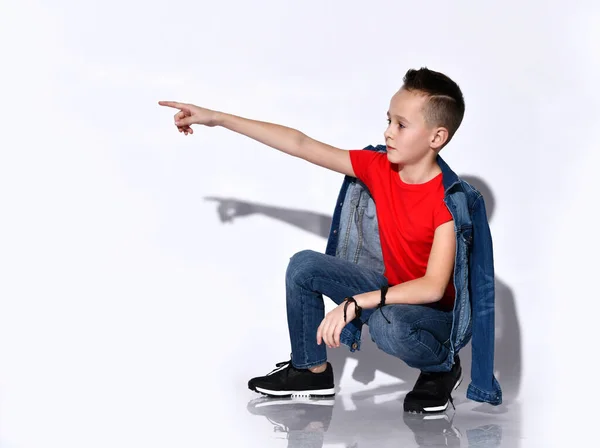 Niño adolescente en camiseta roja, jeans y chaqueta vaquera, zapatillas negras. Señalando algo, poniéndose en cuclillas de lado aislado en blanco —  Fotos de Stock