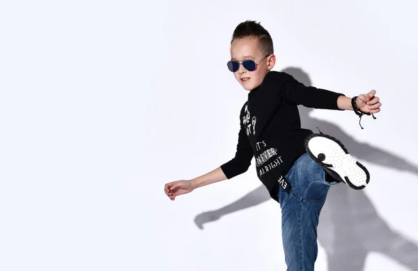 Nastoletnie dziecko w okularach przeciwsłonecznych, czarny sweter i trampki, niebieskie dżinsy, bransoletka. Taniec, pozowanie na białym tle studio — Zdjęcie stockowe