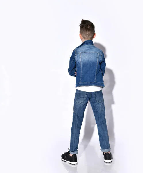 Ragazzo in t-shirt, jeans blu e giacca di jeans. Egli è in piedi dalla schiena alla fotocamera mentre posa isolato su bianco — Foto Stock