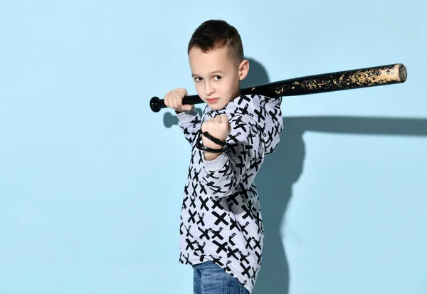 Teenage kluk v mikině a džínách. Ukazuje pěst, drží černou ošuntělou baseballovou pálku nebo hůl, pózuje bokem na modrém pozadí — Stock fotografie