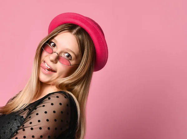 Adolescente femenina en gafas de sol, vestido negro y sombrero rojo. Mostrando su lengua posando de lado sobre fondo rosa. De cerca. — Foto de Stock