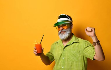 Yeşil güneşlik, gömlek ve güneş gözlüklü sakallı adam. Bir bardak sıkılmış meyve suyu, sıkılmış yumruk, turuncu arka planda poz.