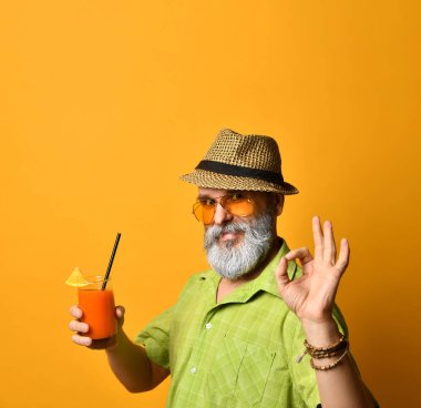 Şapkalı, yeşil gömlekli, güneş gözlüklü sakallı dede. Gülümsüyor, iyiye işaret ediyor, meyve suyu bardağı tutuyor, turuncu arka planda poz veriyor.