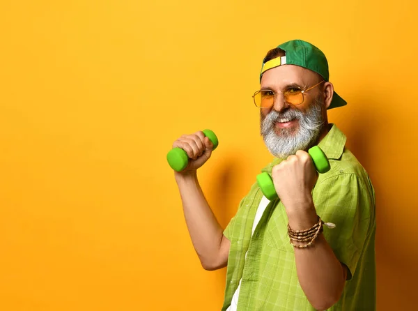 Grandpa in green cap and shirt, white t-shirt, bracelet. He smiling, doing exercises with dumbbells, posing on orange background — Stock Photo, Image