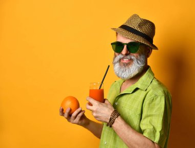 Şapkalı, yeşil gömlekli, güneş gözlüklü yaşlı bir adam. Turuncu arka planda durarak, portakal suyu tutarak ve tüple taze meyve suyu içerek.