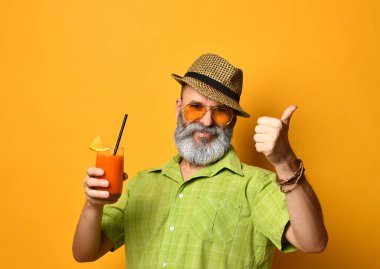 Şapkalı, yeşil gömlekli, güneş gözlüklü sakallı dede. Gülümsüyor, iyiye işaret ediyor, meyve suyu bardağı tutuyor, turuncu arka planda poz veriyor.