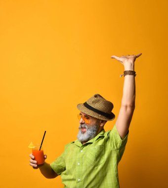 Sakallı, şapkalı, yeşil gömlekli, güneş gözlüklü bir erkek. Dans eder gibi elini kaldırdı. Elinde meyve suyu bardağıyla turuncu arka planda poz verdi.