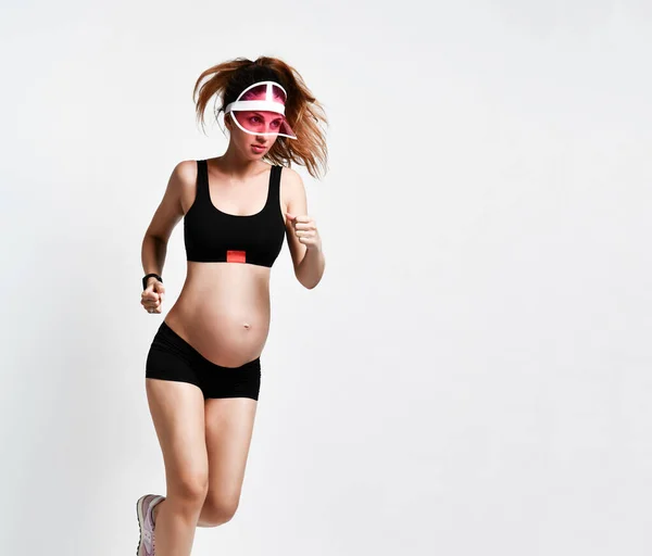 Kobieta w ciąży w okularach przeciwsłonecznych, czarne spodenki sportowe i top, różowe trampki. Uśmiecha się podczas joggingu na białym. Pełna długość — Zdjęcie stockowe