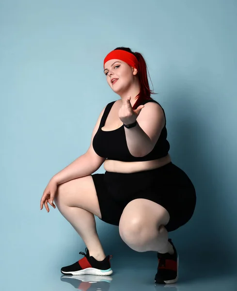 Model imbiru z nadwagą w czerwonej opasce, czarny top, szorty, trampki. Squatting pozowanie bokiem na tle niebieskiego studio — Zdjęcie stockowe