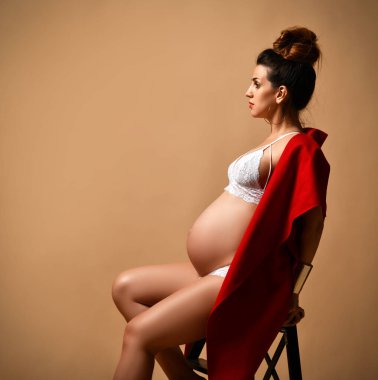 Küpeli esmer kadın, beyaz iç çamaşırı ve kırmızı şal. Bej arka planda poz verirken yan koltukta oturuyor. Yakın plan.