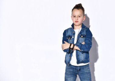 Tişörtlü, kot pantolonlu, kot ceketli, siyah bilezik takan genç bir çocuk. Gücenmiş görünüyor, elleri katlanmış, bembeyaz poz veriyor.