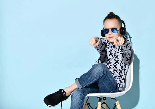 Дитина-підліток в навушниках, сонцезахисних окулярах, синіх джинсах, светрі, кросівки. Підборіддя підборіддя за кулаком, сидячи на стільці. Синій фон — стокове фото