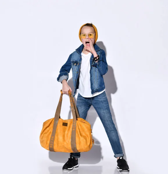 オレンジの旅行バッグ、帽子、サングラス、 Tシャツ、ブルーデニムスーツ、ブラックスニーカーを持つ10代の子供。彼は白に隔離されたポーズ — ストック写真