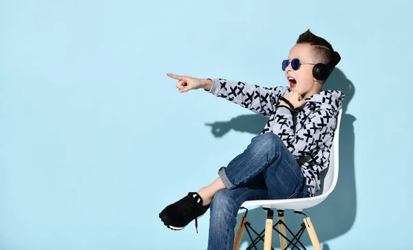 Nastolatek w słuchawkach, okularach przeciwsłonecznych i ubraniach. Wskazując na coś, siedząc na białym krześle na niebieskim tle — Zdjęcie stockowe