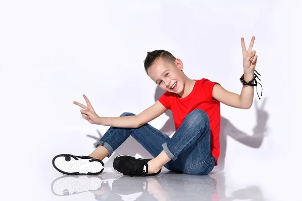Tonårskille i röd t-shirt, blå jeans, svarta sneakers. Sitter på golvet, ler, visar segertecken, poserar isolerad på vitt — Stockfoto
