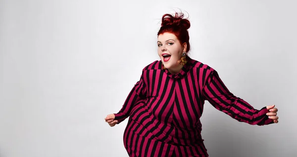 Obese redhead lady in striped dress and earrings. She reaching her hand to you while posing sideways isolated on white background — Stockfoto