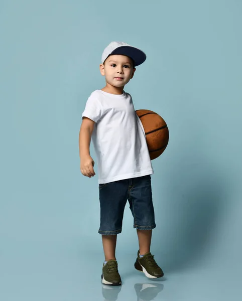 Ragazzo in t-shirt, berretto, pantaloncini e sneakers. Tenendo palla da basket, sorridente, mostrando tre dita, posa su sfondo blu — Foto Stock