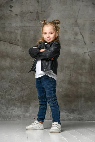 Klein schattig lachend blond meisje in stijlvolle rock stijl zwart lederen jas, jeans en witte sneakers staan in zelfverzekerde pose — Stockfoto