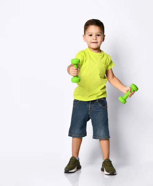 Mały chłopiec w żółtej koszulce, jeansowe szorty, trampki khaki. Uśmiechnięta, trzymająca zielone hantle, pozująca na białym tle — Zdjęcie stockowe