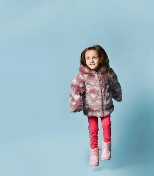 Klein meisje gekleed in roze nepbontjas, broek en laarzen. Ze glimlacht, gebarentaal, poseert op een blauwe achtergrond. Totale lengte — Stockfoto