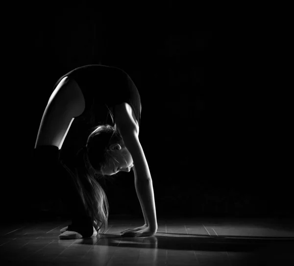 Giovane ginnasta ragazza in corpo nero sport e calzature speciali in piedi in posa ponte e mantenere l'equilibrio su sfondo scuro — Foto Stock