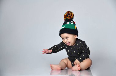 Siyah elbiseli, şapkalı, timsah izli, yalınayak bir çocuk. Gülümsüyor, beyaz zeminde tek başına oturuyor. Kapat.