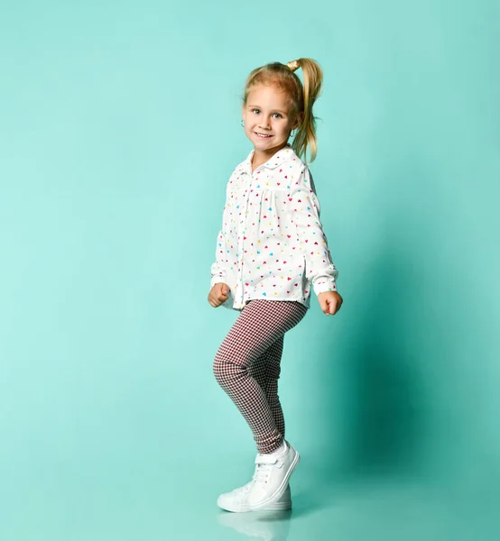 Kleine blonde jongen in shirt met hartafdruk, geruite broek, witte sneakers. Lachend, dansend op een blauwe achtergrond. Totale lengte — Stockfoto