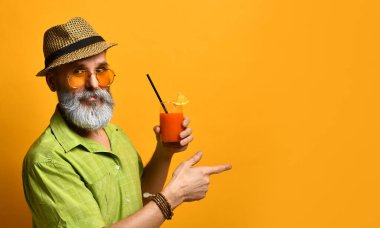 Büyükbaba şapkalı, yeşil gömlekli, güneş gözlüklü. Bıyık bükme, elinde taze sıkılmış meyve suyu bardağı, turuncu arka planda poz verme.