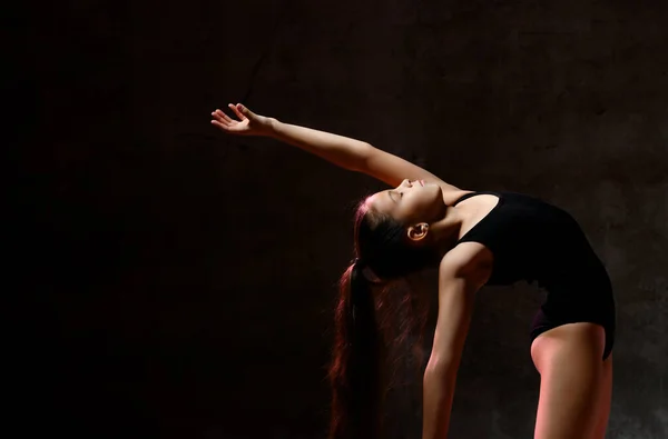 Chica joven gimnasta en cuerpo deportivo negro y calzado especial haciendo pose gimnástica sobre fondo oscuro — Foto de Stock