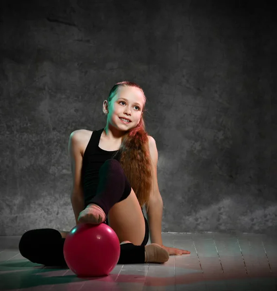 Jong lachend meisje turner in zwarte sport lichaam en bovendelen zitten op de vloer en aanraken roze gymnastiek bal met been — Stockfoto