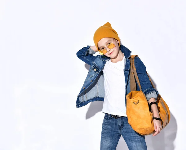 Porträtt Ett tonårigt barn med en orange resväska, klädd i en hatt, solglasögon, en T-shirt, en blå jeansdräkt, svarta sneakers och ett armband. — Stockfoto