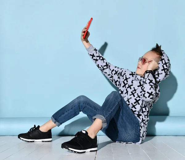 Enfant en lunettes de soleil, jeans et sweat à capuche, baskets. Prendre selfie par smartphone rouge, assis latéralement sur le sol blanc. Fond bleu — Photo